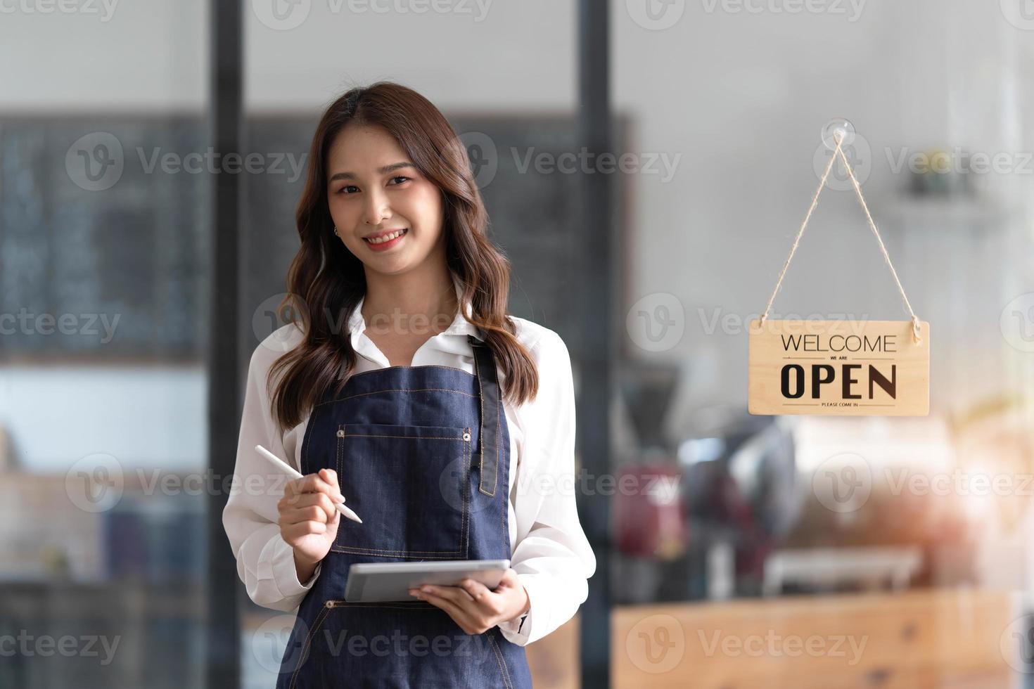 mooi Aziatisch jong barista vrouw in schort Holding tablet en staand in voorkant van de deur van cafe met Open teken bord. bedrijf eigenaar opstarten concept. foto