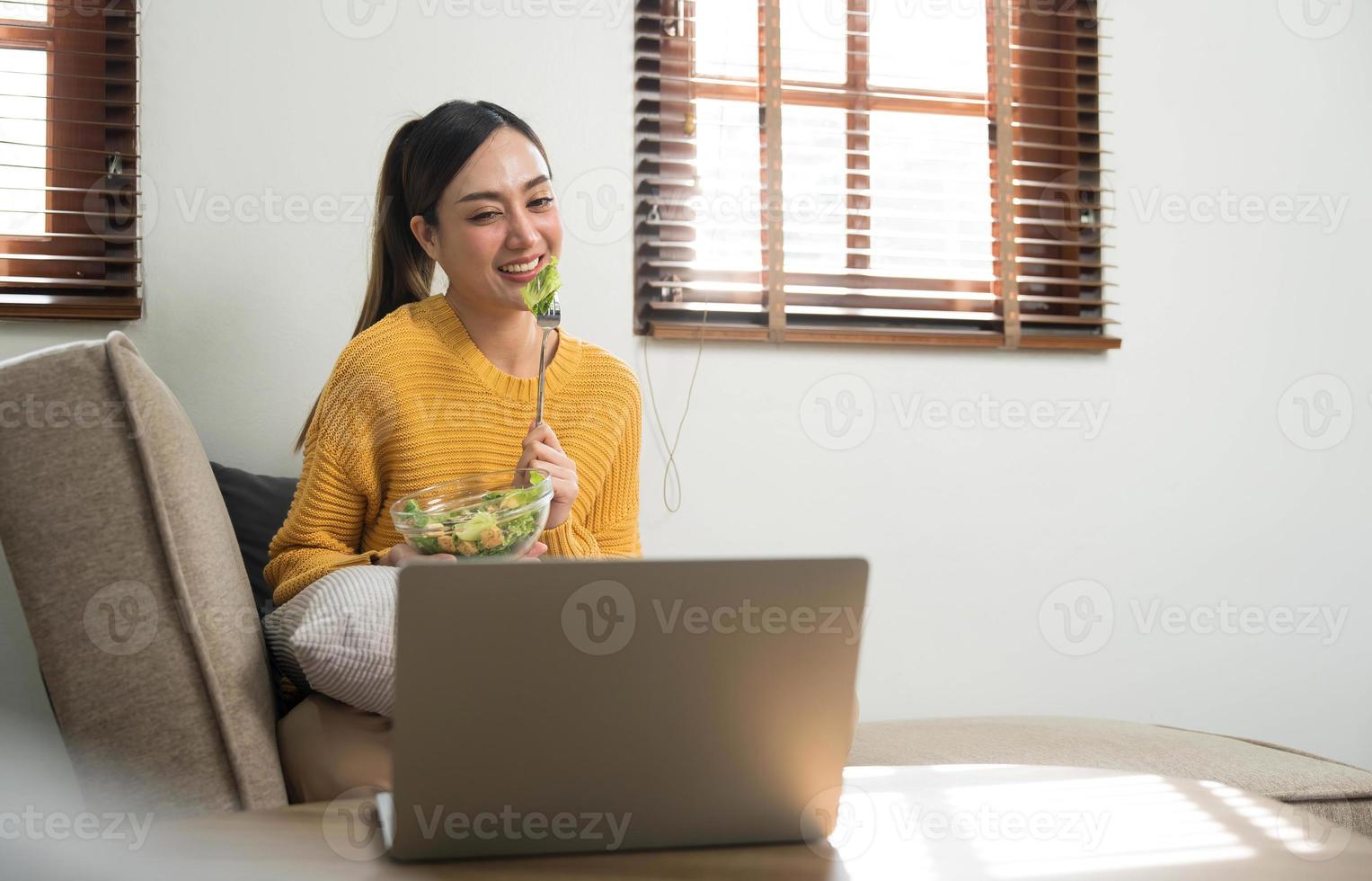 mensen kom tot rust Bij huis en welzijn levensstijl. jong volwassen Aziatisch vrouw aan het eten salade en gebruik makend van laptop computer voor aan het kijken online film Aan internetten. foto