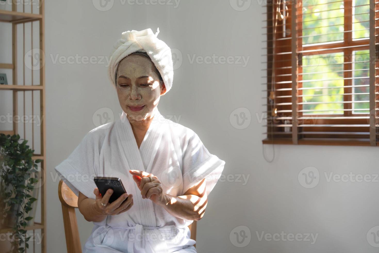 mooi volwassen Aziatisch dame in badjas met gelaats hydraterend of anti veroudering klei masker Aan haar gezicht is gebruik makend van haar mobiel telefoon in de leven kamer. foto