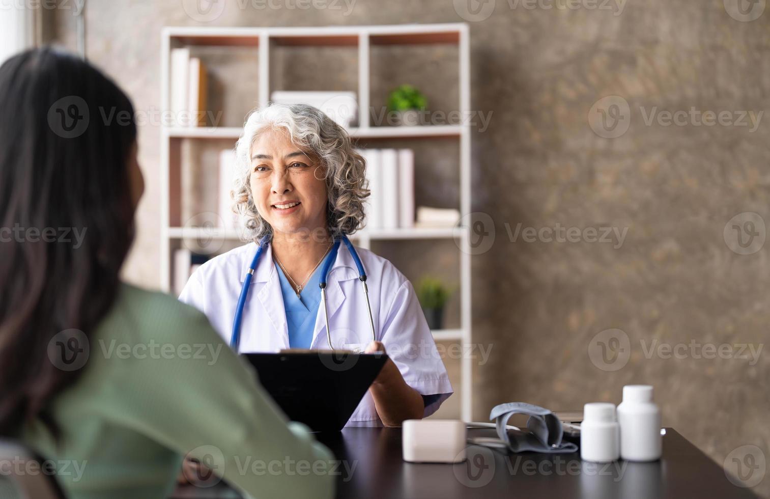 vrouw senior dokter is lezing medisch geschiedenis van vrouw geduldig en sprekend met haar gedurende overleg in een Gezondheid kliniek. arts in laboratorium jas zittend achter een laptop in ziekenhuis kantoor. foto