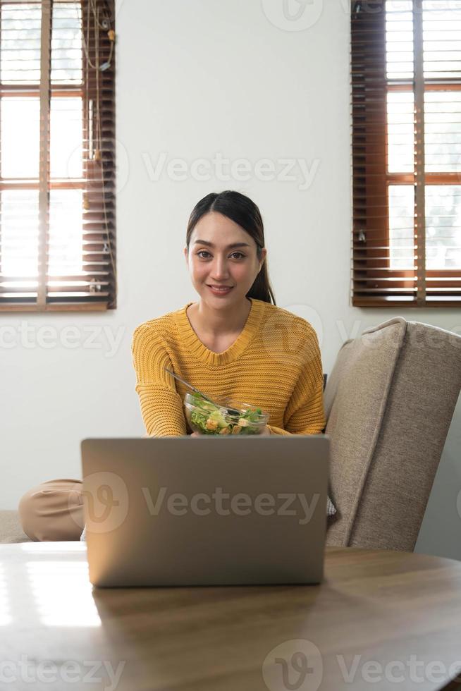mensen kom tot rust Bij huis en welzijn levensstijl. jong volwassen Aziatisch vrouw aan het eten salade en gebruik makend van laptop computer voor aan het kijken online film Aan internetten. foto