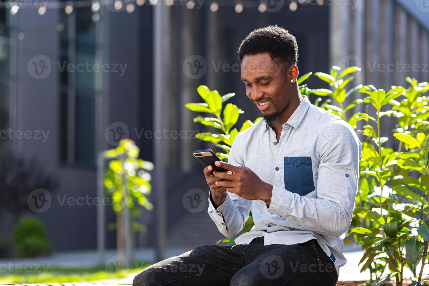 vrolijk Afrikaanse Amerikaans vent gebruik makend van mobiel telefoon zittend Aan bank in stad park buitenshuis foto