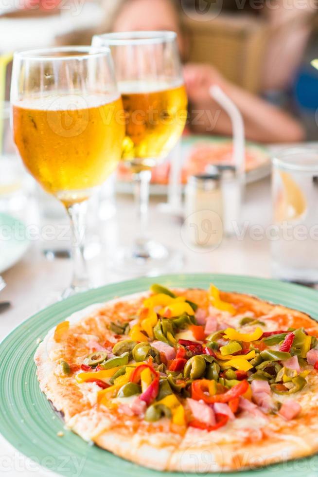 pizza met Mozzarella kaas, olijf, vers tomaat en pesto saus. geserveerd Bij restaurant tafel met twee bieren foto