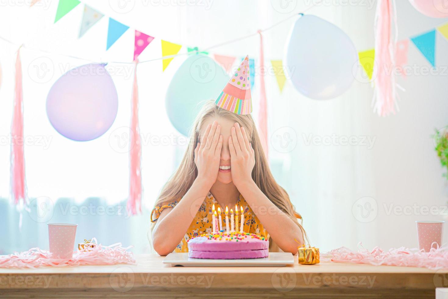Kaukasisch meisje is dromerig glimlachen en op zoek Bij verjaardag regenboog taart. feestelijk kleurrijk achtergrond met ballonnen. verjaardag partij en wensen concept. foto