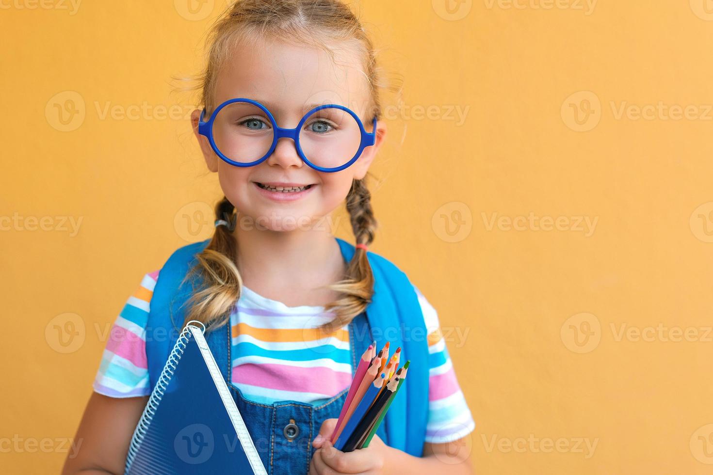 portret glimlachen school- meisje in bril met school- rugzak houdt gekleurde potloden en blauw notebook. klaar voor school. terug naar school. geïsoleerd Aan een geel achtergrond kopiëren ruimte. reclame uitverkoop foto