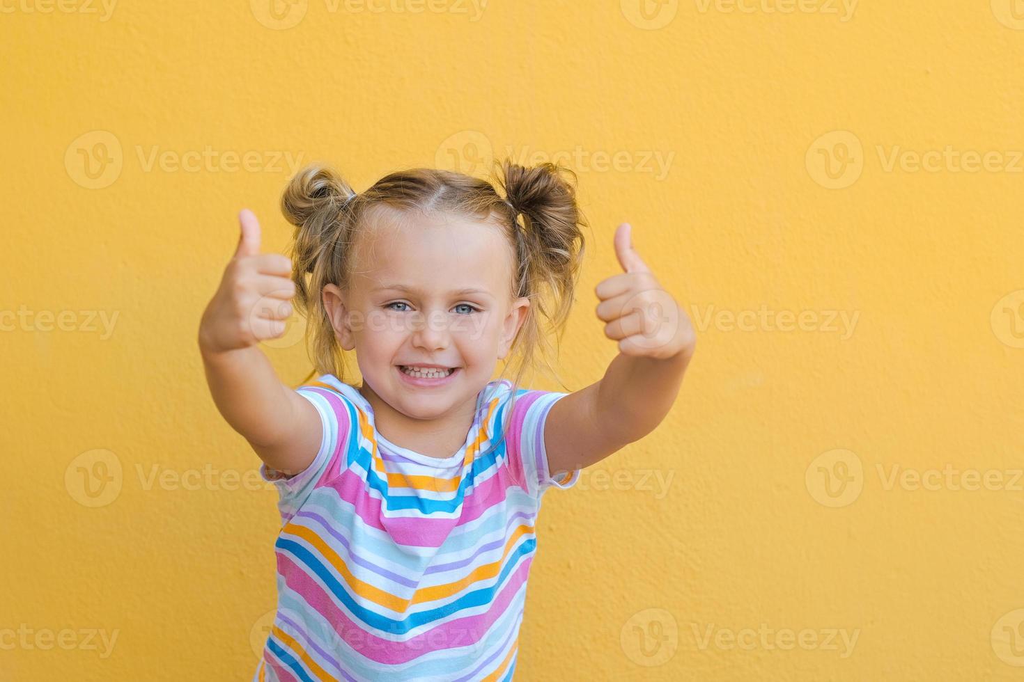 portret gelukkig verrassing weinig kind meisje in gekleurde t-shirt tonen punt vingers duimen omhoog handen glimlachen adviseren geïsoleerd over- geel kleur achtergrond foto