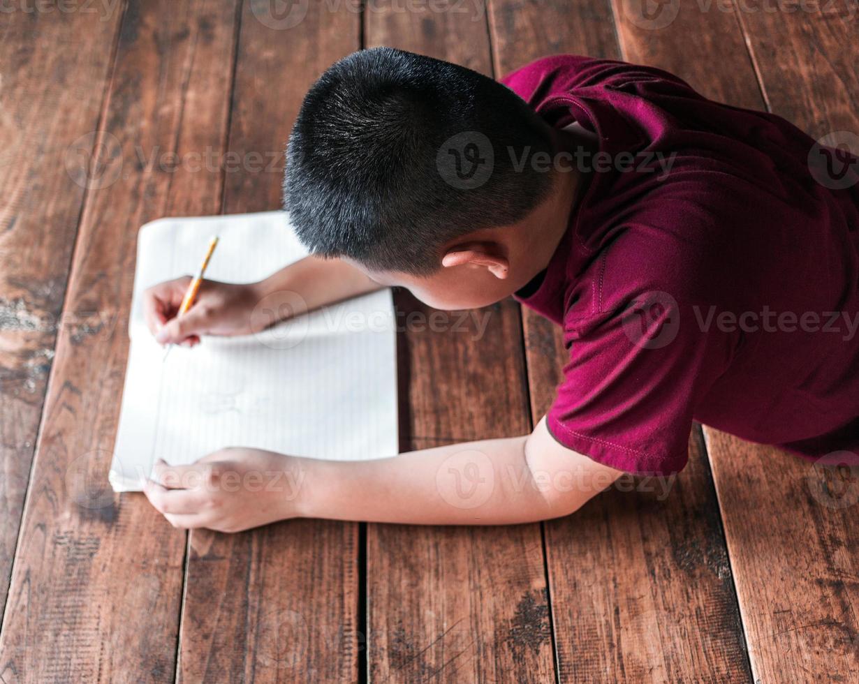 jongen zittend Aan de houten verdieping schrijven een boek of aan het doen huiswerk Bij huis. top visie van kind schrijven boek foto