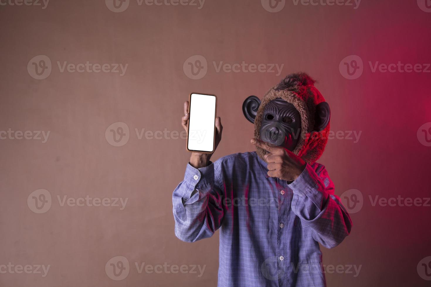 Aziatisch Mens vervelend blauw lang overhemd met aap masker tonen telefoon, foto