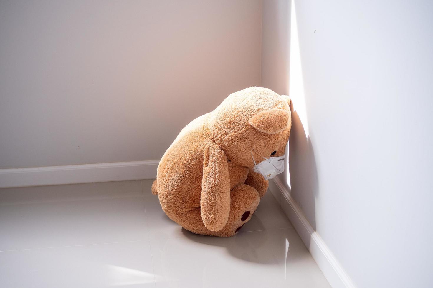 de teddy beer is vervelend een masker, zittend en boog naar beneden in de hoek van de kamer. depressie, verdrietig, teleurstelling of ziekte foto