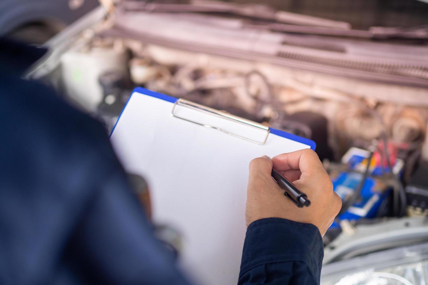 handen van jong professioneel monteur in uniform schrijven Aan klembord tegen auto in Open kap Bij de reparatie garage. onderhoud zie. auto onderhoud en onderhoud foto