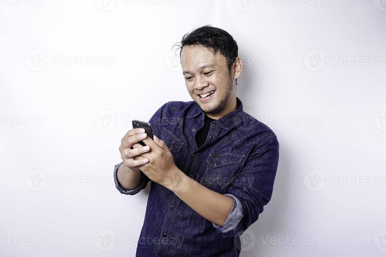 opgewonden Aziatisch Mens vervelend blauw overhemd glimlachen terwijl Holding zijn telefoon, geïsoleerd door wit achtergrond foto