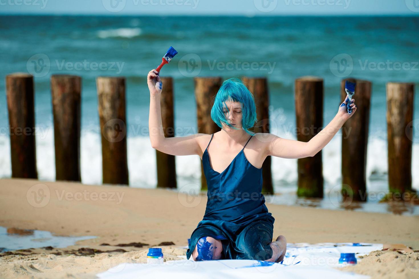 artistiek blauwharig vrouw prestatie artiest besmeurd met blauw gouache verven zittend Aan strand foto