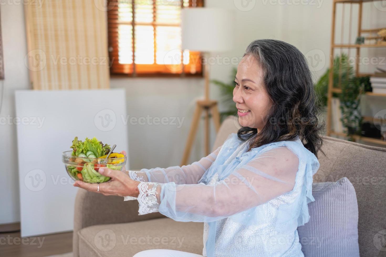 Aziatisch senior vrouw zittend aan het eten groente salade en gezond voedsel en aan het eten gelukkig Aan de sofa in de huis voor een gezond lichaam. gezond voedsel concept foto