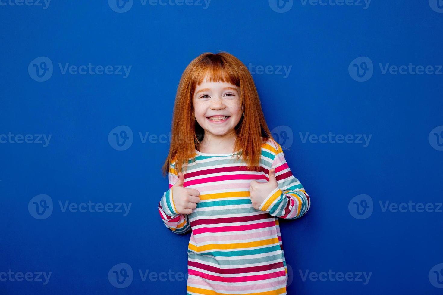 weinig meisje met rood haar- in een veelkleurig glimlach staat Aan een blauw achtergrond foto