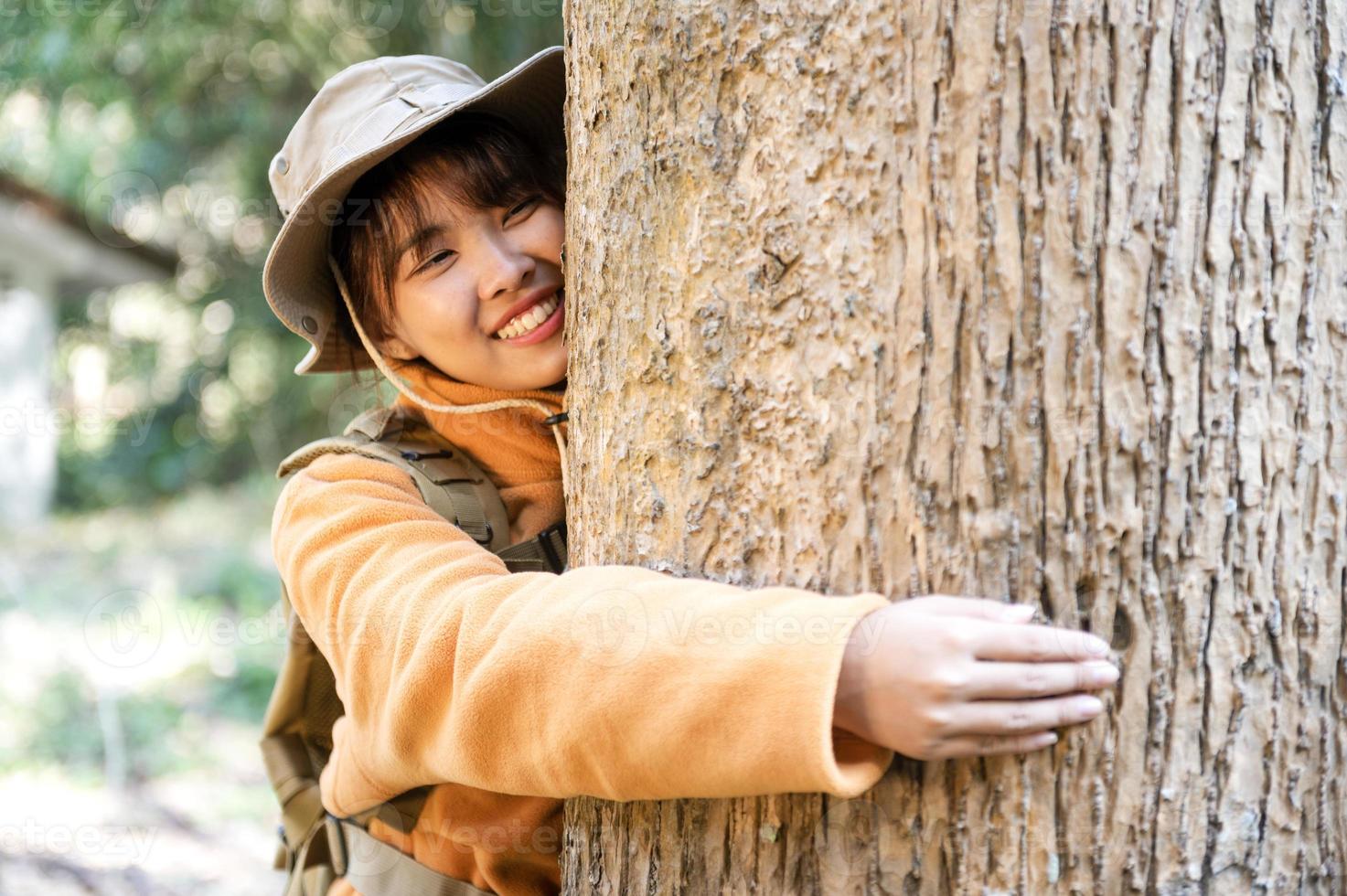 jong toerist vrouw in een geel jas knuffelen een boom in de Woud van eco liefde op zoek omhoog Bij de boomtoppen jong Aziatisch vrouw onderzoeken een groot ecologisch boom foto