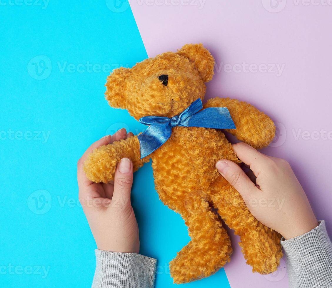 twee vrouw handen houden een klein bruin speelgoed- teddy beer Aan een blauw Purper achtergrond foto