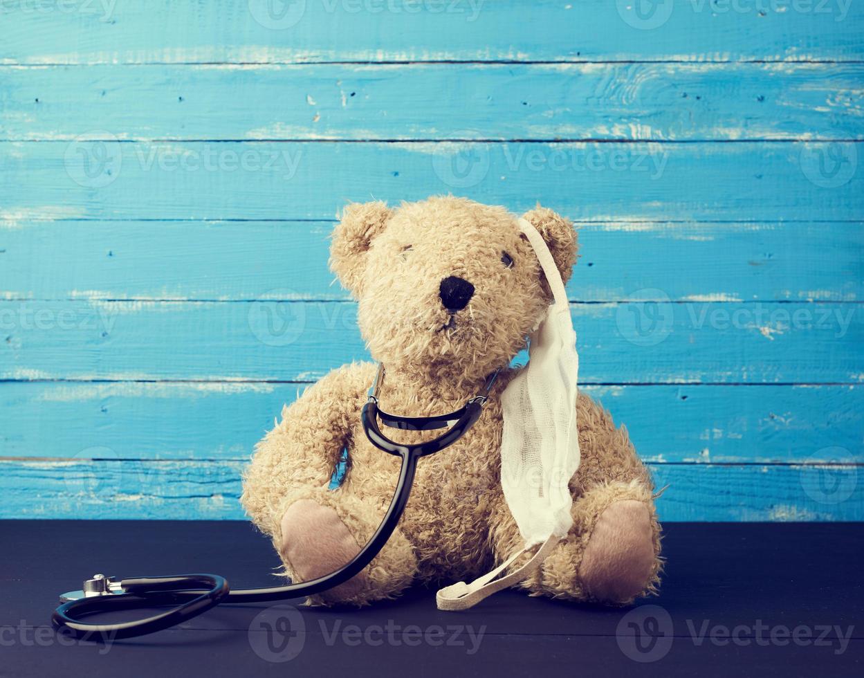 teddy beer is zittend in een wit medisch masker, zwart stethoscoop is hangende Aan zijn nek foto