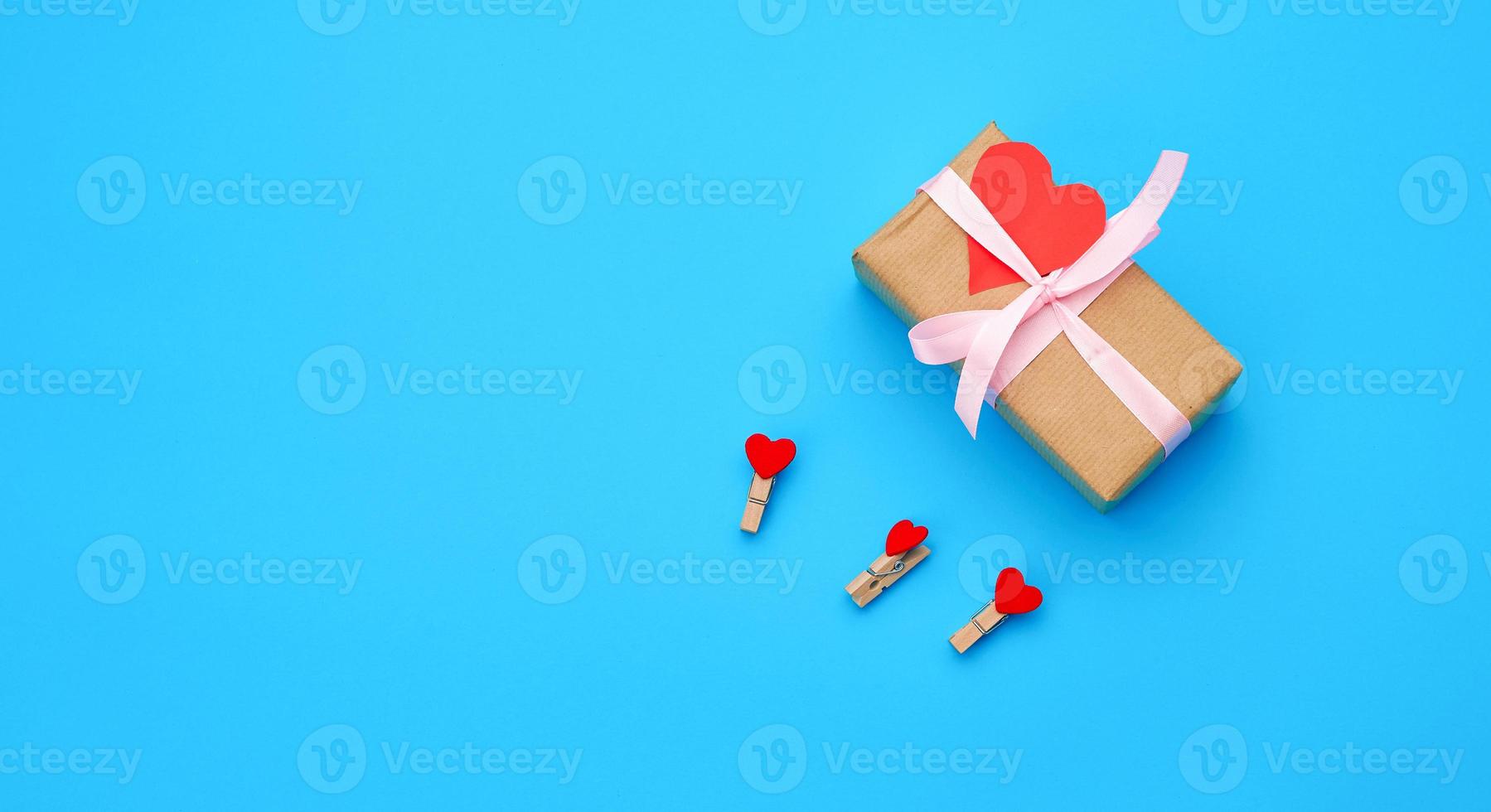doos is verpakt in bruin papier en gebonden met een rood dun zijde lint Aan een blauw achtergrond, rood besnoeiing uit papier harten foto