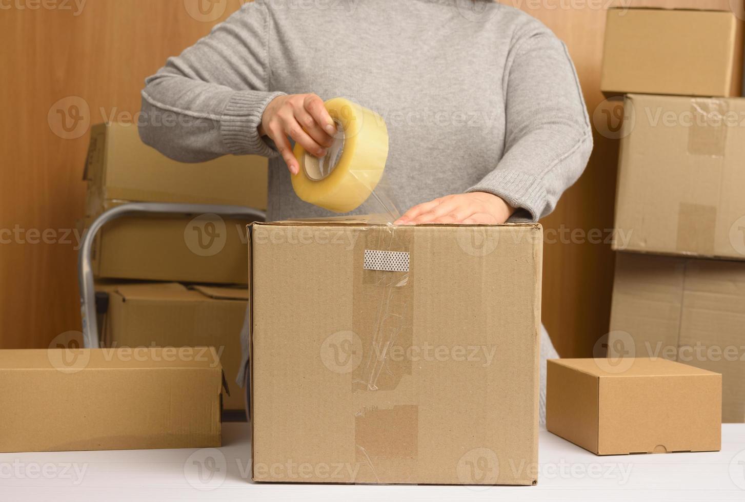 vrouw in een grijs trui houdt een rollen van kanaal plakband en pakketten bruin karton dozen Aan een wit tafel, achter een stack van dozen foto