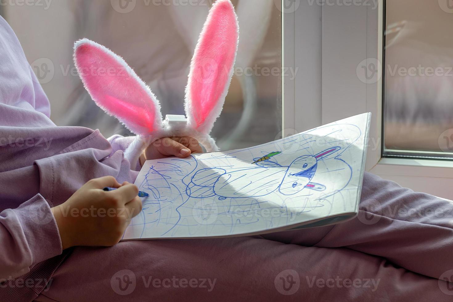 een schoolmeisje meisje met konijn oren zit Aan de venster en trekt een konijn in haar album, voorbereidingen treffen voor Pasen foto