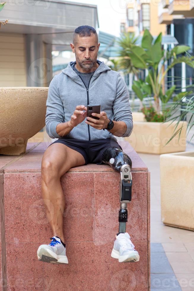 sportman met prothetisch been zittend Aan blok en gebruik makend van mobiele telefoon foto
