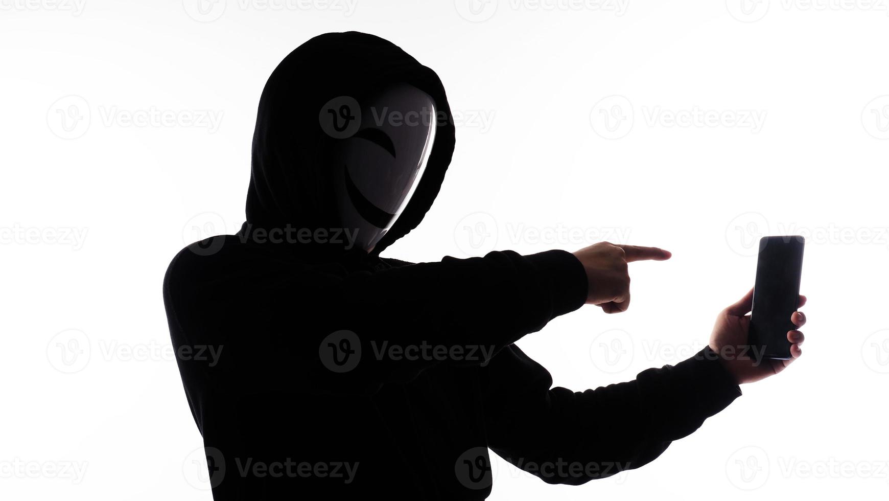 hacker anoniem en gezicht masker met smartphone in hand. Mens in zwart kap overhemd Holding en gebruik makend van mobiel telefoon Aan wit achtergrond. staan voor cyber misdrijf gegevens hacken of stelen persoonlijk gegevens concept foto