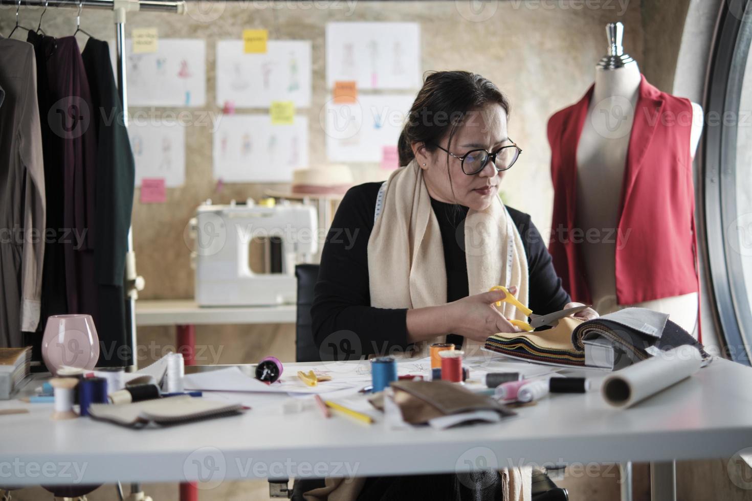 Aziatisch middelbare leeftijd vrouw mode ontwerper werken in studio, snijdend en kiezen kleding stof en draad kleuren met tekening schetsen voor jurk ontwerp collecties. professioneel winkel kleermaker ondernemer. foto