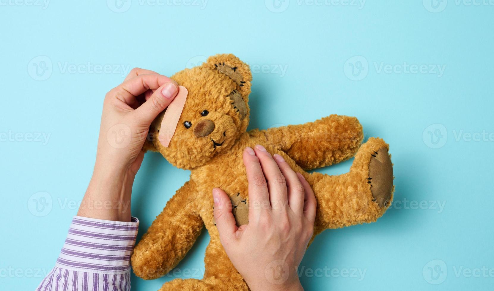 vrouw hand- houdt een bruin teddy beer en lijmen een medisch Zelfklevend gips Aan een blauw achtergrond, tram behandeling foto