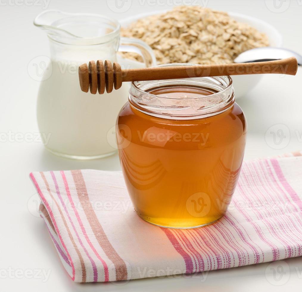 honing in een glas transparant pot en een houten stok Aan een wit tafel, achter een karaf met melk en havermout foto