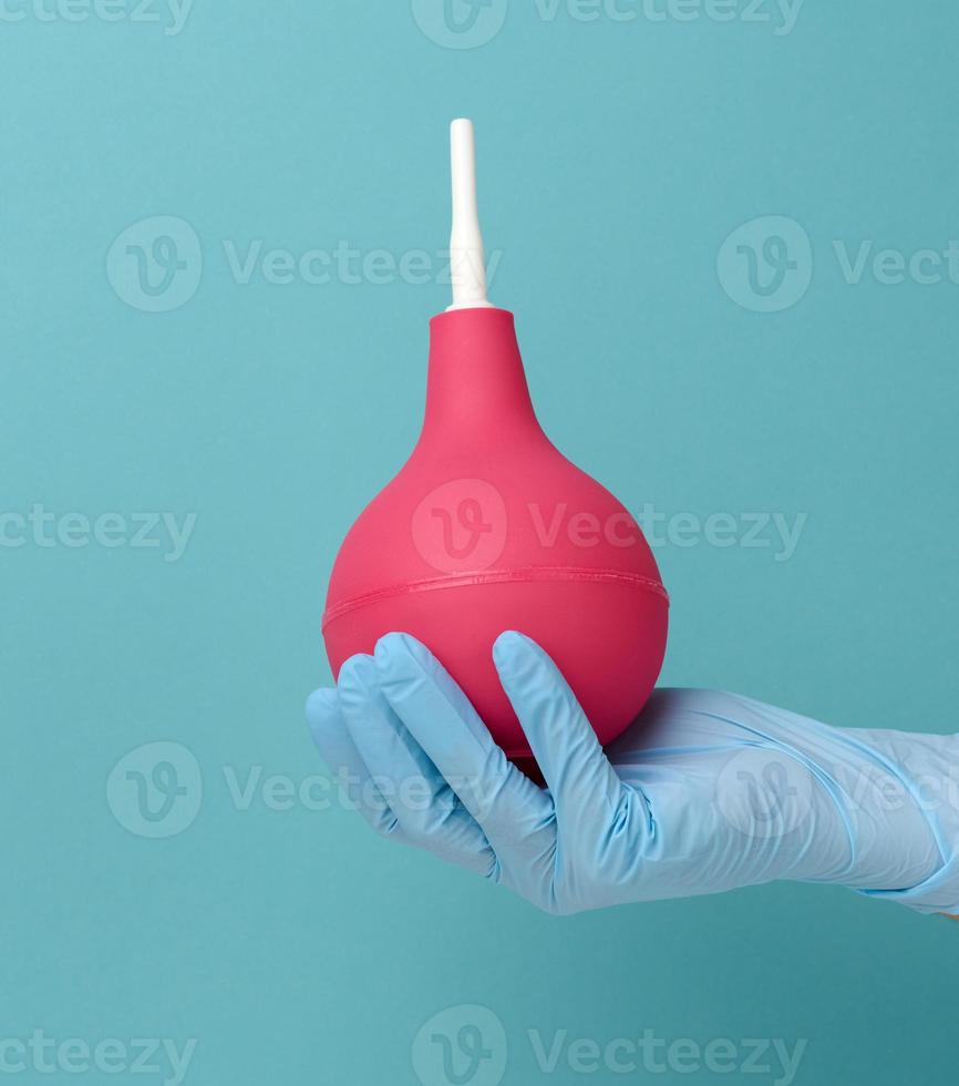een hand- in een blauw medisch handschoen houdt een roze rubber klysma Aan een blauw achtergrond foto