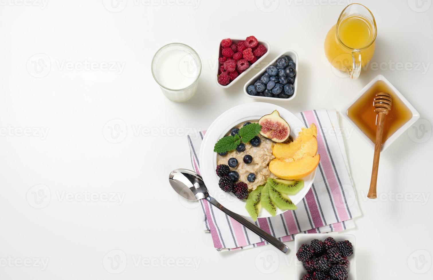 melk, bord met havermout pap en fruit, vers geperst sap in een transparant glas karaf, honing in een kom Aan een wit tafel. gezond ontbijt foto