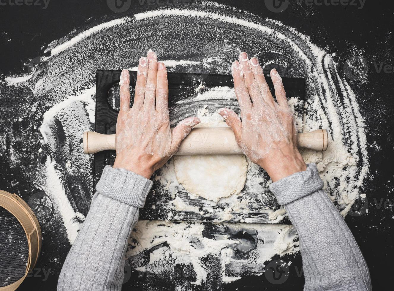 twee vrouw handen rollen uit wit tarwe meel deeg met een houten rollend pin Aan een zwart tafel, top visie foto