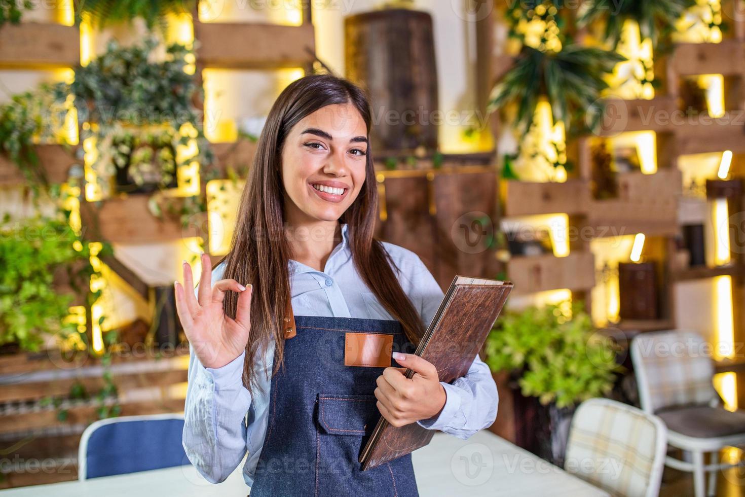 mooi vrouw serveerster glimlachen in de restaurant Holding een menu en tonen OK teken. foto