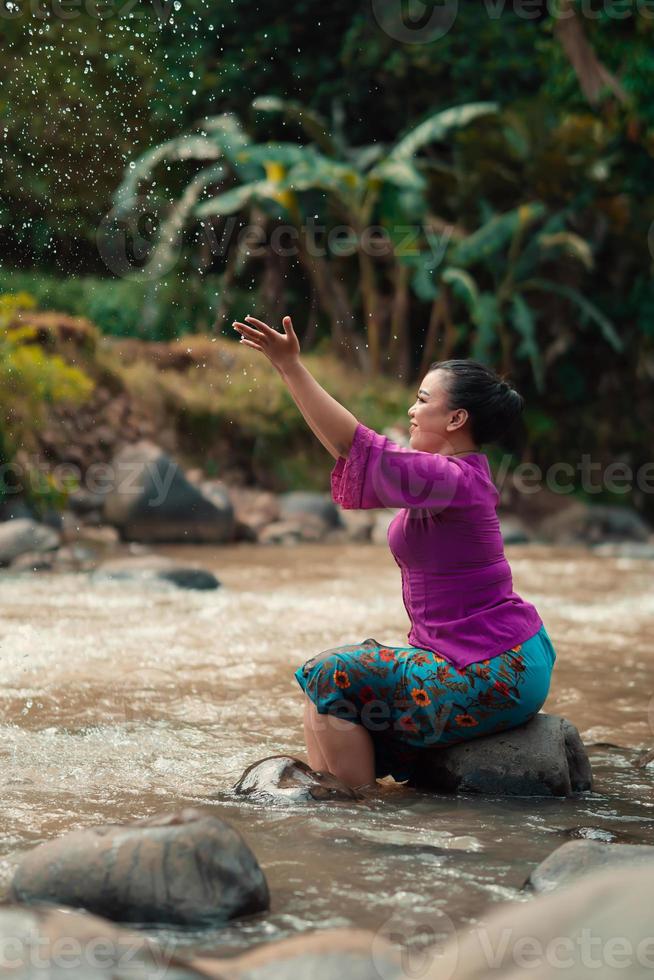 Indonesisch vrouw zittend Aan een klein rots en spelen met de water terwijl haar jurk kreeg nat van de rivier- foto