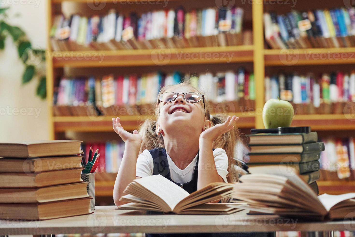 gevoel gelukkig. schattig weinig meisje met vlechten is in de bibliotheek. appel Aan de boeken foto