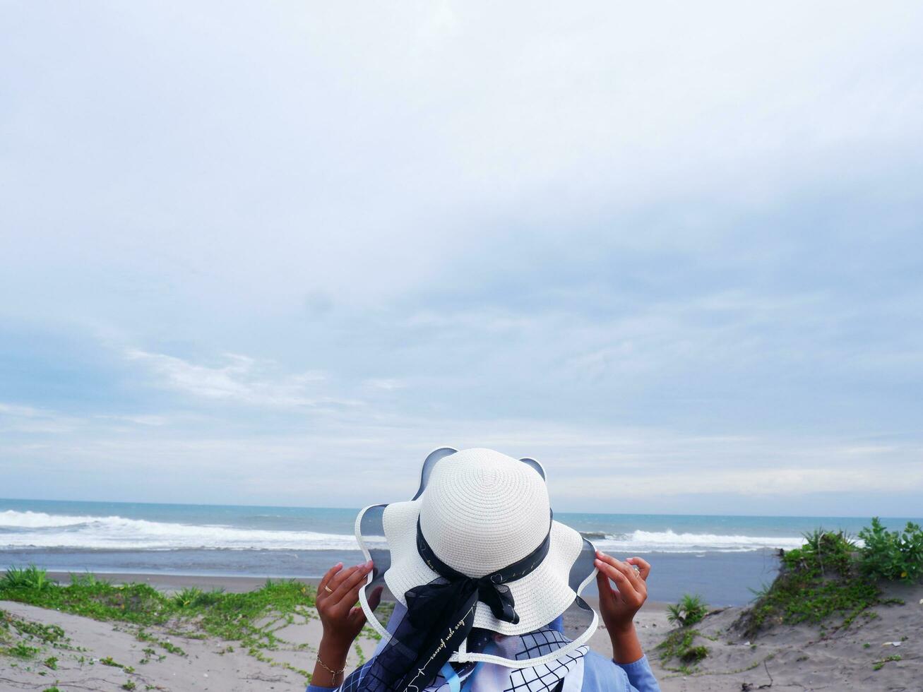 terug van de vrouw in de hoed Aan de tropisch strand wie is op zoek Bij de lucht en de zee terwijl Holding haar hoed foto