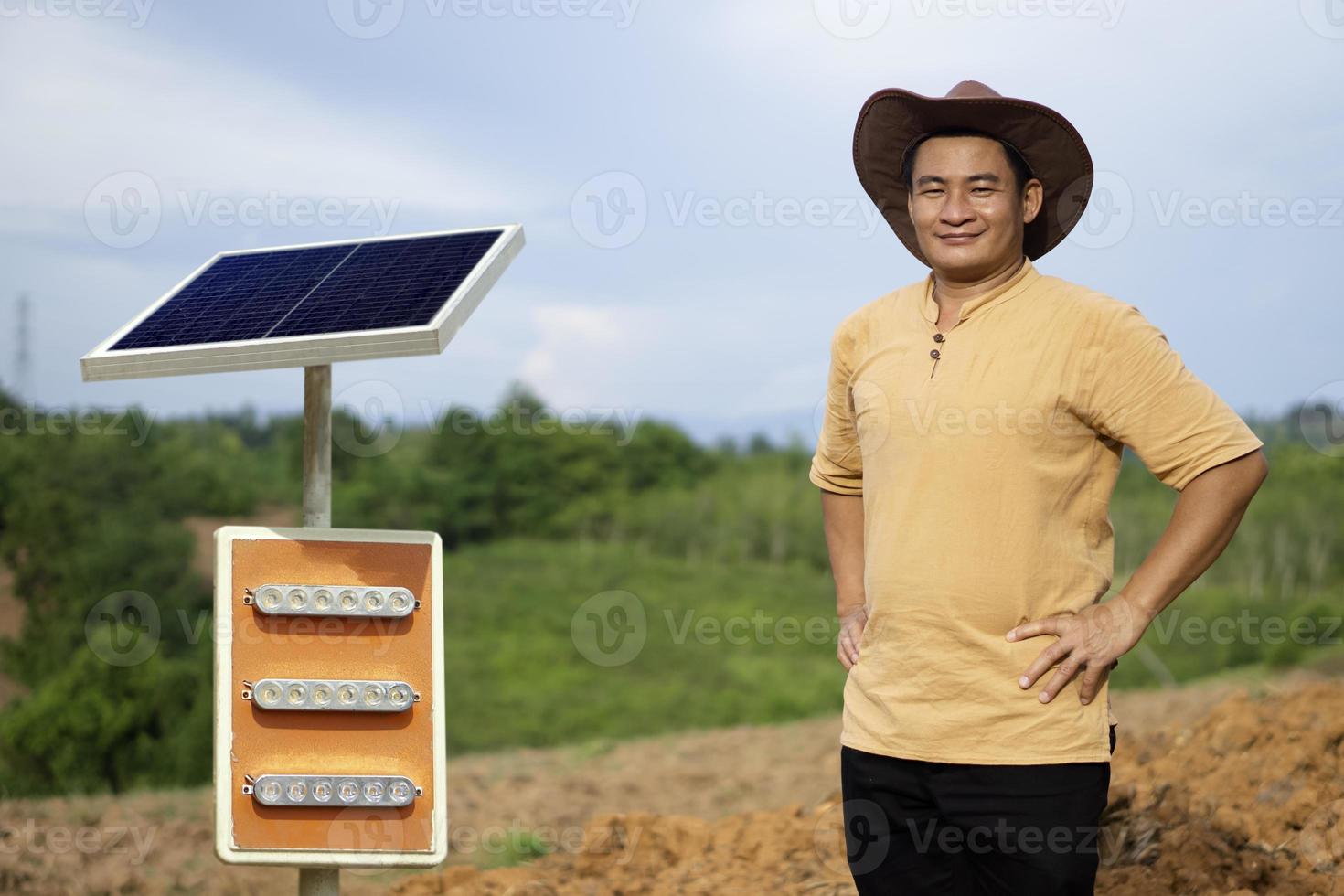 Aziatisch mannetje boer, draagt hoed, bruin shirt, zetten handen Aan heupen, staat naast mini zonne- paneel. concept, groen technologie landbouw. hernieuwbaar energie, natuurlijk stroom. milieuvriendelijk levensstijl. foto