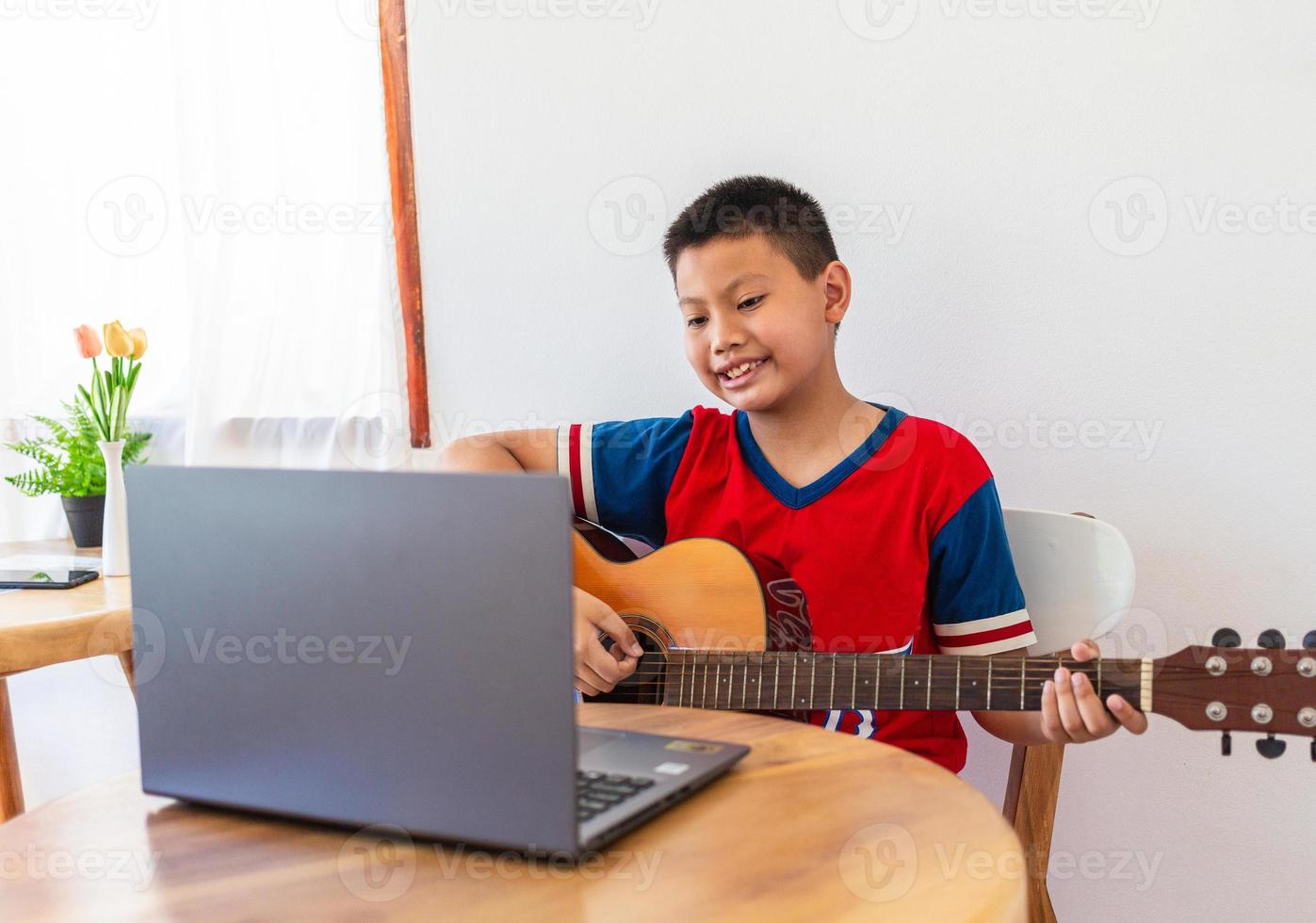 de verhaal van een jongen aan het kijken een notitieboekje computer terwijl voorbereidingen treffen naar praktijk spelen gitaar Bij huis. jongens nemen klassiek gitaar lessen online. foto