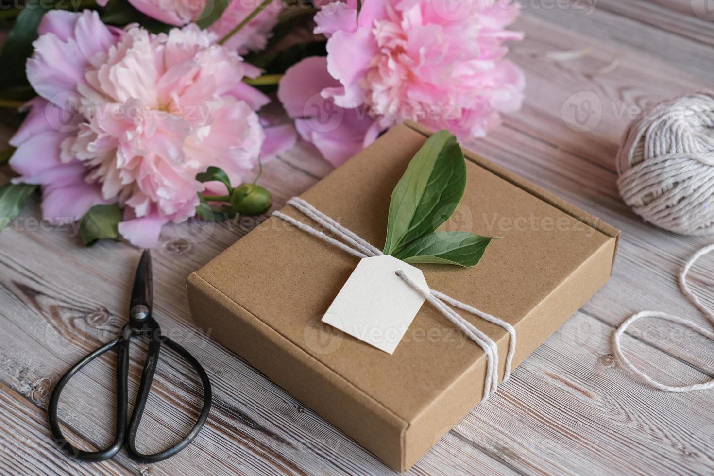 bespotten omhoog label, kraft geschenk doos versierd met bloemen pioenrozen. voorbereiding van cadeaus voor moeder dag, verjaardag, vrouwen vakantie. geschenk omhulsel idee foto