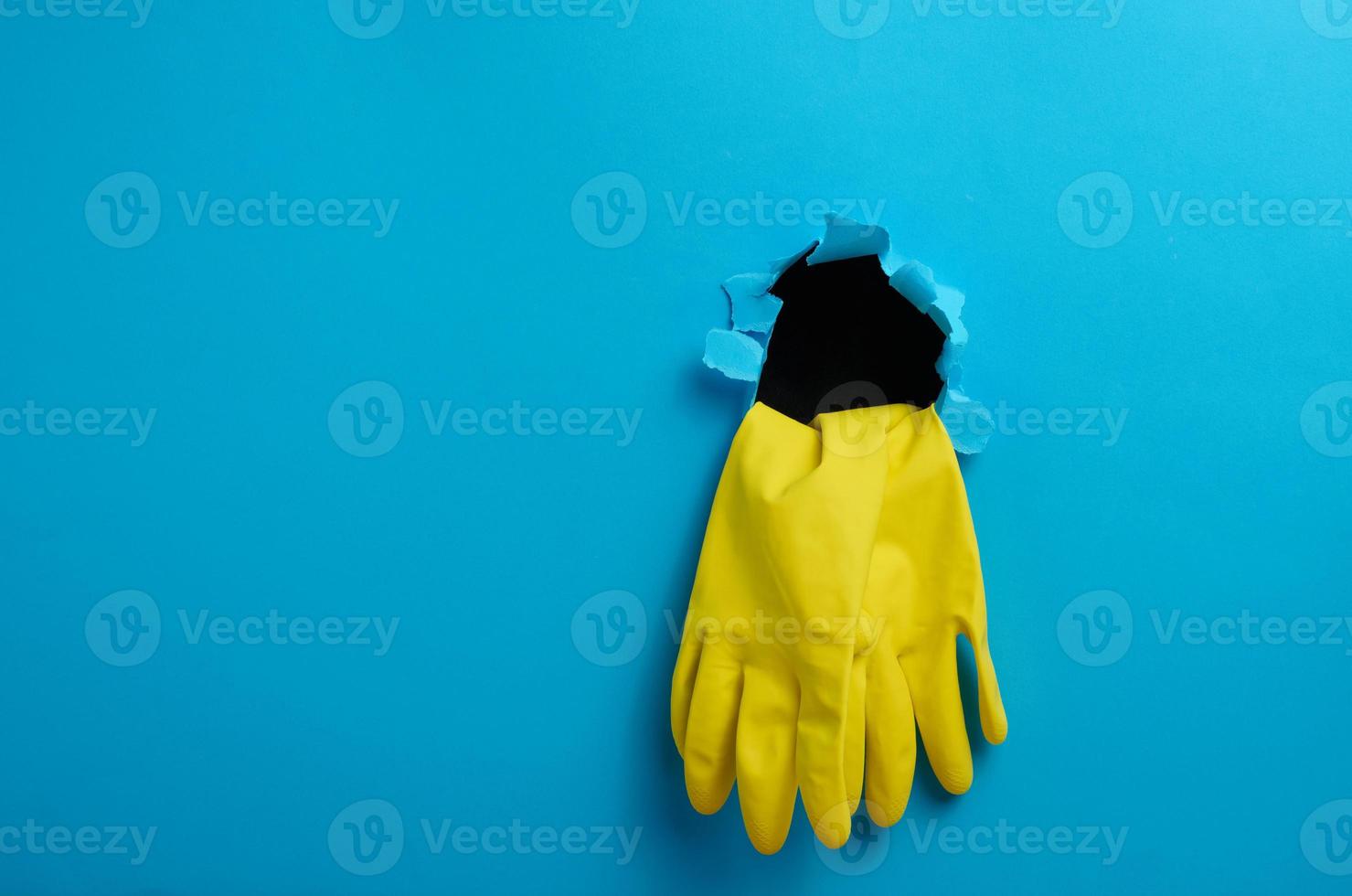 een paar- van geel latex huis schoonmaak handschoenen plakken uit van de gescheurd gat van de blauw papier achtergrond foto