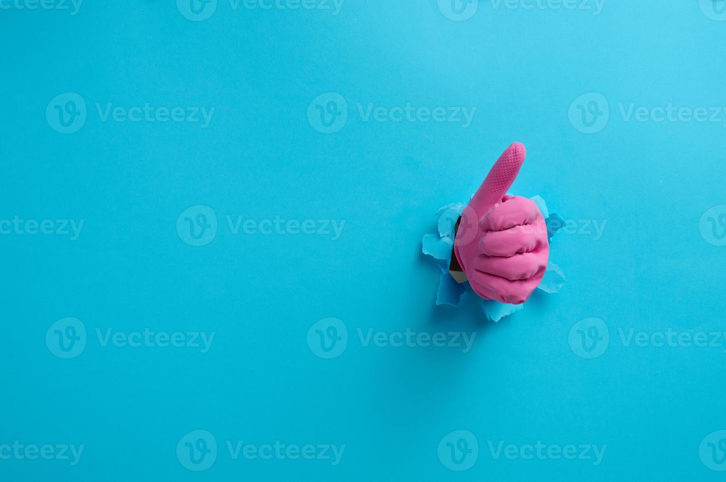 een hand- in een roze huishouden rubber handschoen stokjes uit van de gescheurd gat en shows een gebaar Oke, duim omhoog. blauw achtergrond foto