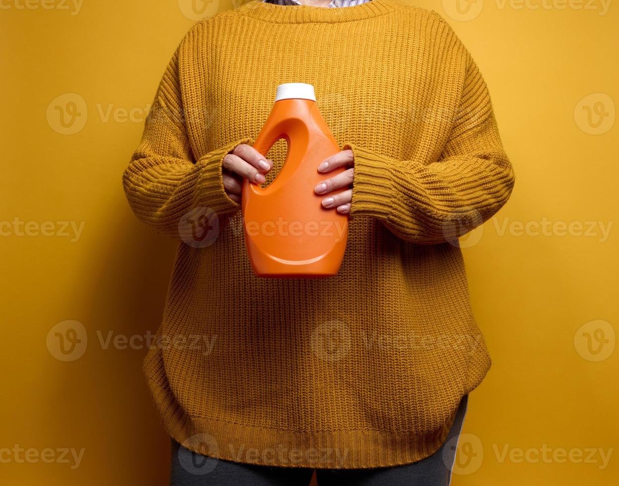 vrouw in een gebreid trui houdt een oranje plastic fles met vloeistof het wassen gel. wasserij en klusjes, geel achtergrond foto