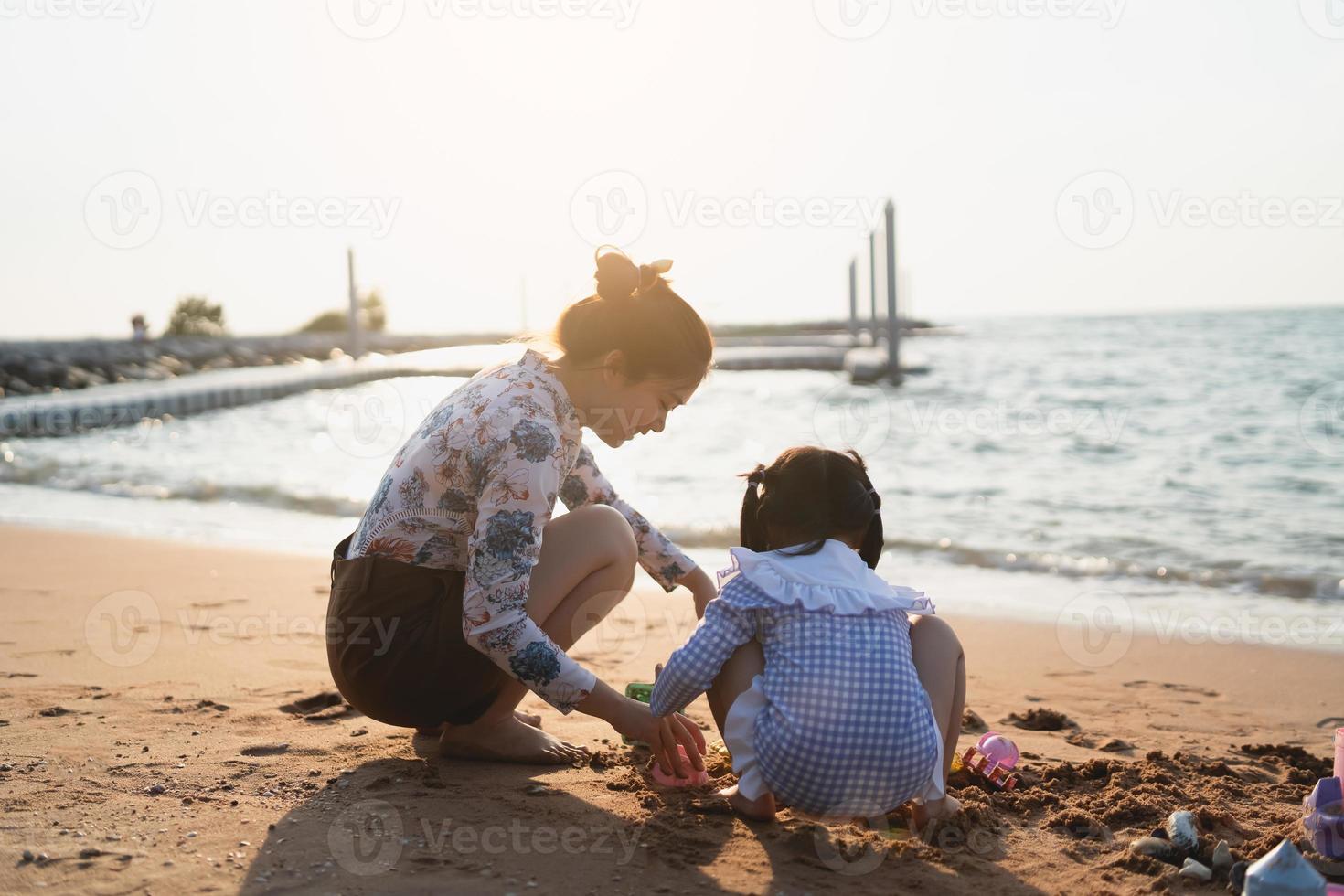 Aziatisch schattig weinig meisje en haar moeder spelen of maken zand kasteel of graven met zand Aan tropisch strand. kinderen met mooi zee, zand blauw lucht. gelukkig kinderen Aan vakanties kust Aan de strand. foto