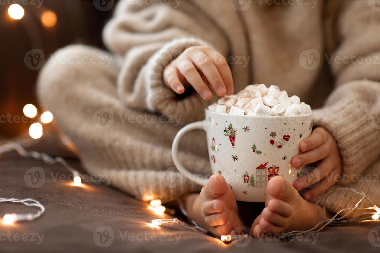 kind kaal voeten en handen houden kop heet cacao marshmallows dichtbij omhoog slinger lichten.flaffy wazig warm gebreid beige trui. Kerstmis concept, vakantie.blij nieuw jaar.kind meisje zittend Aan sofa huis foto