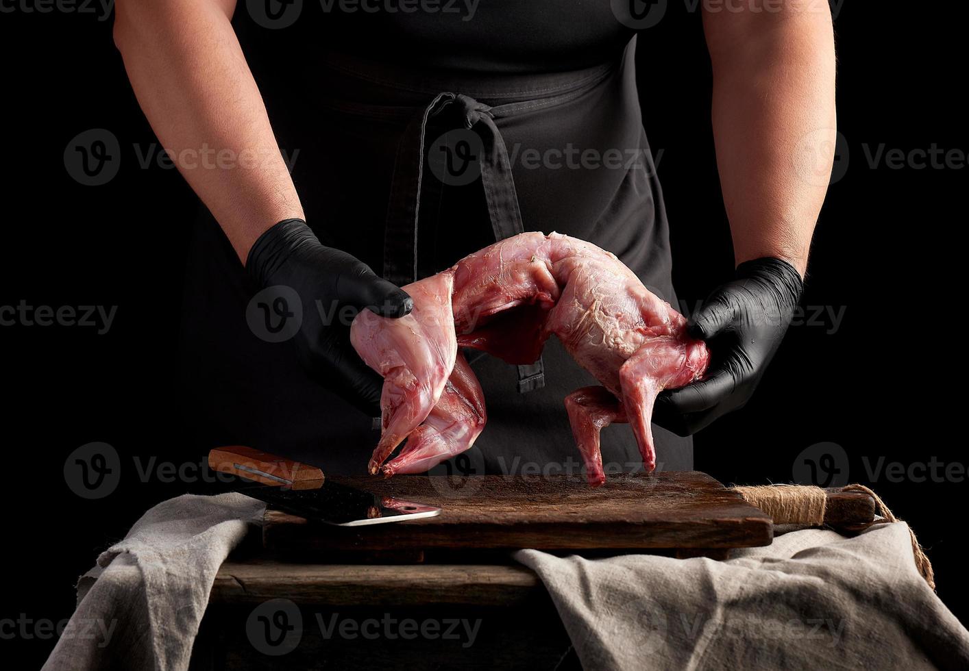 chef in zwart latex handschoenen houdt een geheel konijn karkas over- een bruin snijdend bord, vlees Koken werkwijze foto