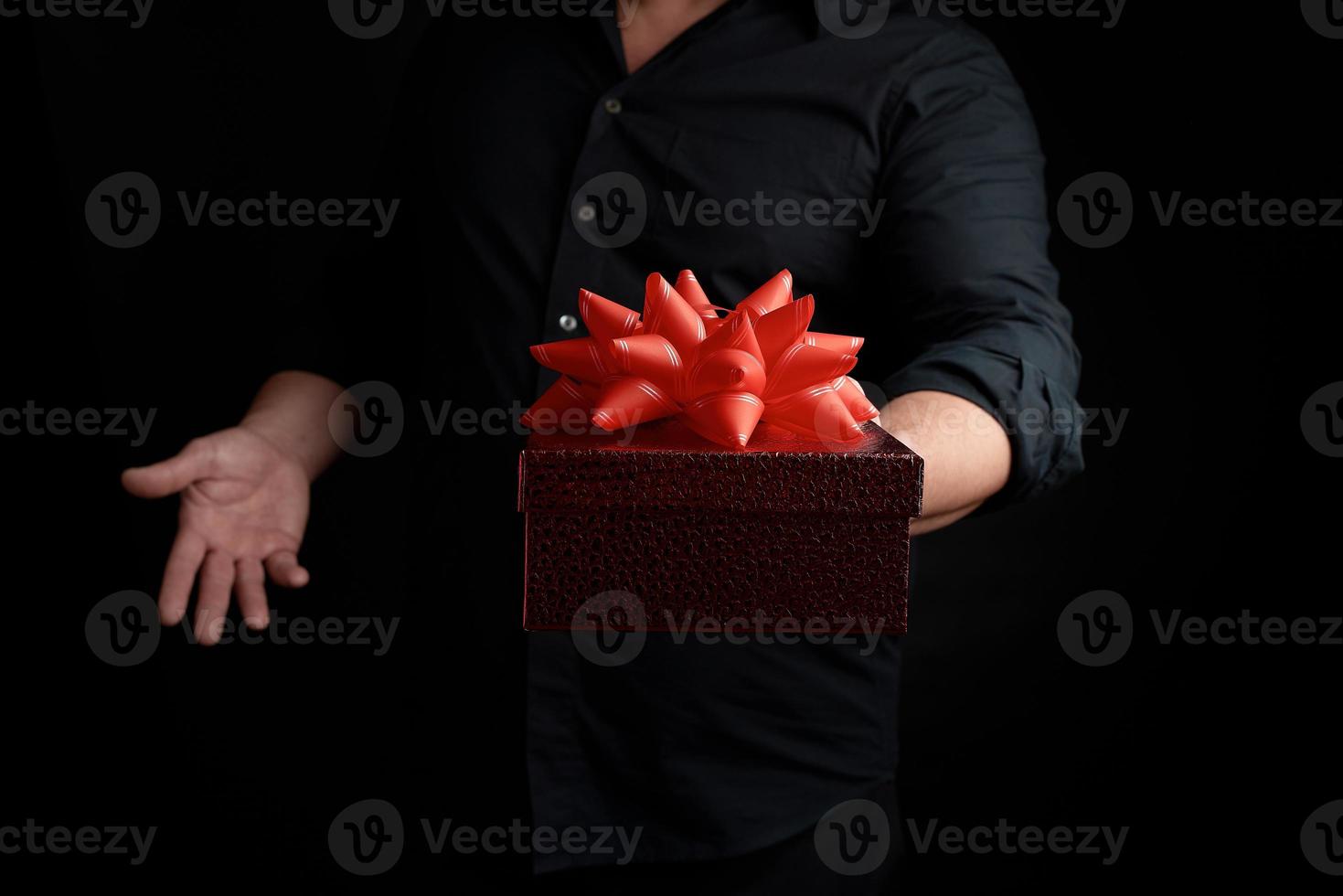 volwassen Mens in een zwart overhemd houdt een rood plein doos met een geknoopt boog Aan een donker achtergrond foto