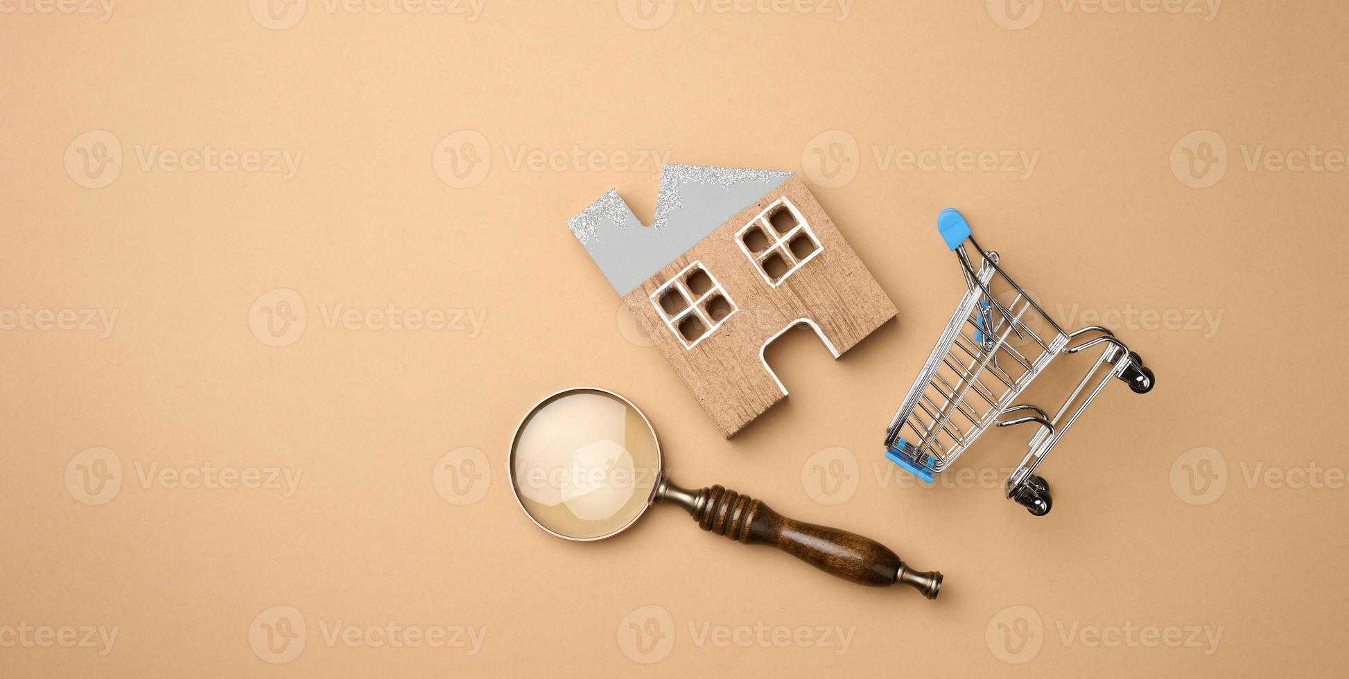 model- van een houten huis, een miniatuur boodschappen doen kar en een vergroten glas Aan een licht bruin achtergrond. huis zoeken concept voor huur, aankoop, hypotheek foto