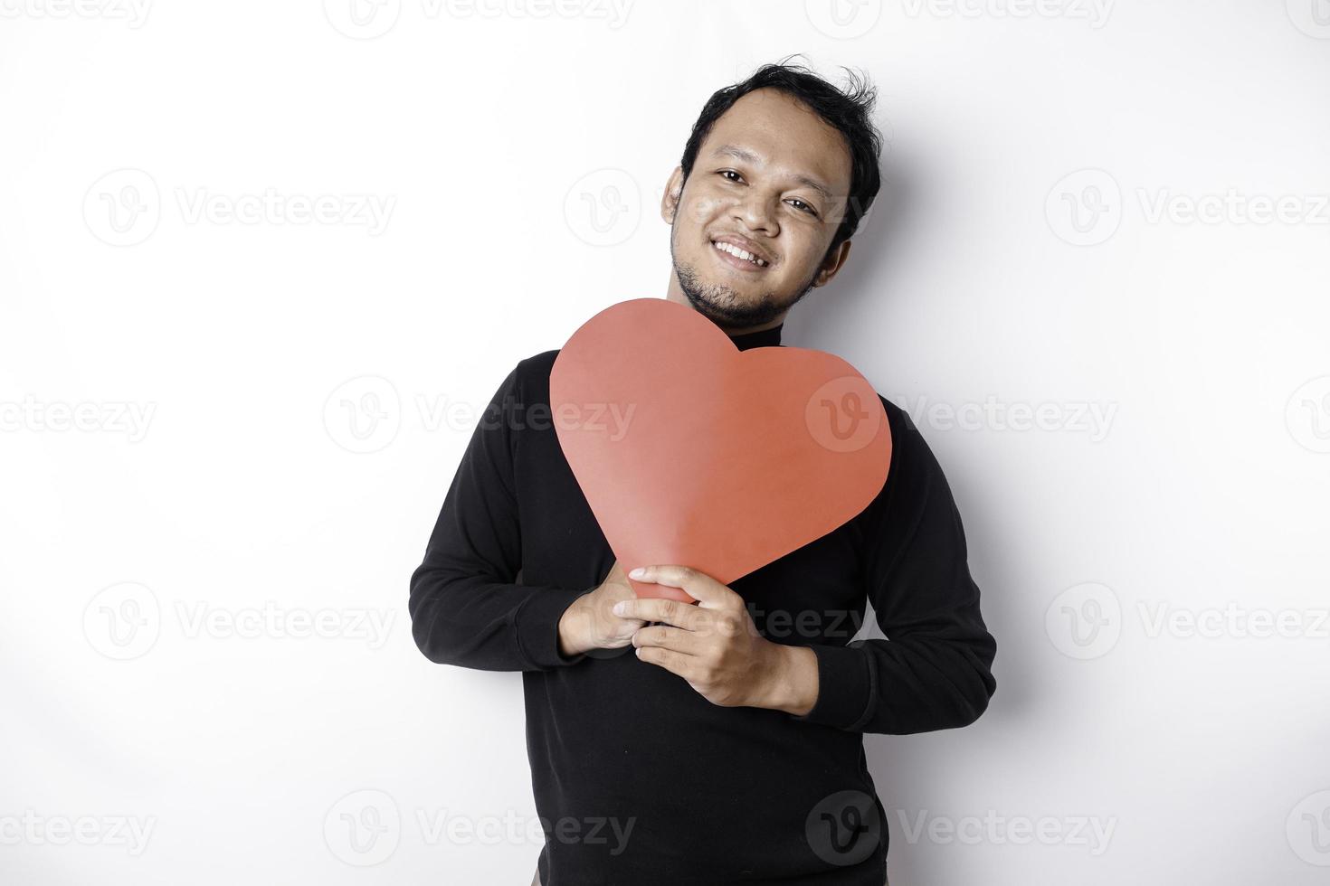 een portret van een gelukkig Aziatisch Mens vervelend een zwart shirt, Holding een rood hartvormig papier geïsoleerd door wit achtergrond foto