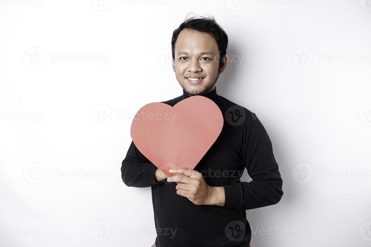 een portret van een gelukkig Aziatisch Mens vervelend een zwart shirt, Holding een rood hartvormig papier geïsoleerd door wit achtergrond foto