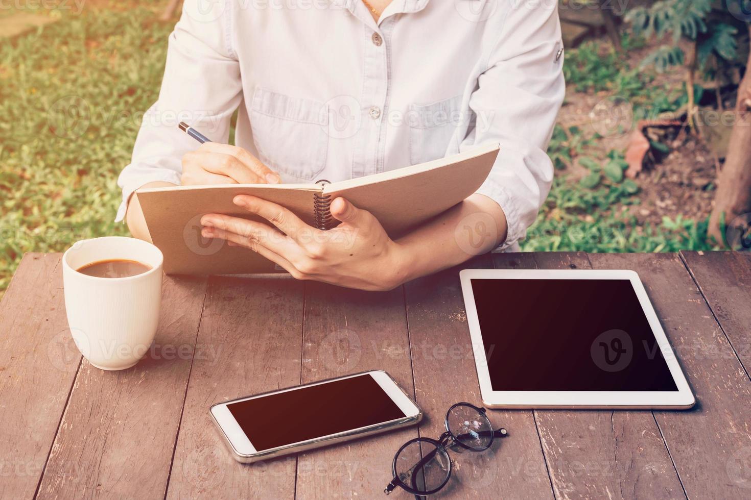 vrouw hand- schrijven notitieboekje en telefoon, tablet Aan tafel in tuin Bij koffie winkel met wijnoogst afgezwakt. foto